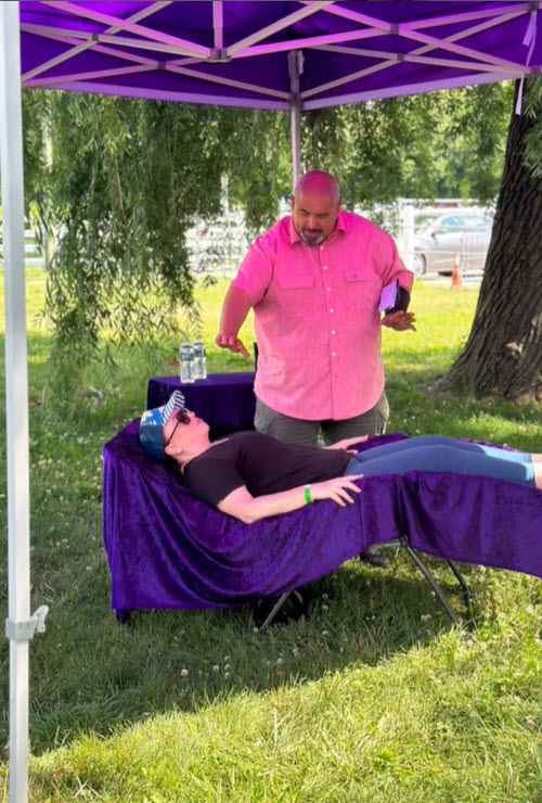 A person with a pink button down shirt standing over another person on an outdoor lounge chair draped with purple fabric.