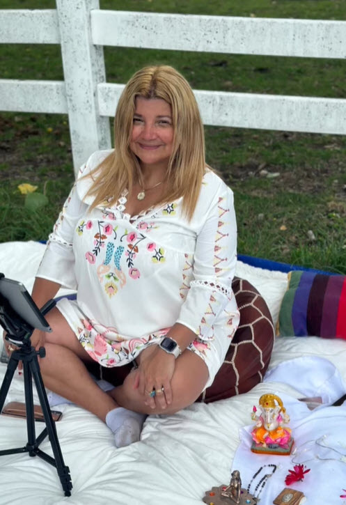 A person wearing a white blouse with colored designs sitting on a comforter outside preparing for a guided healing meditation.